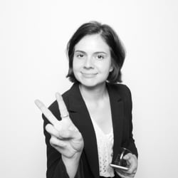 A photo of a smiling person with chin-length brown hair, wearing a blazer and flashing a peace sign to the camera.